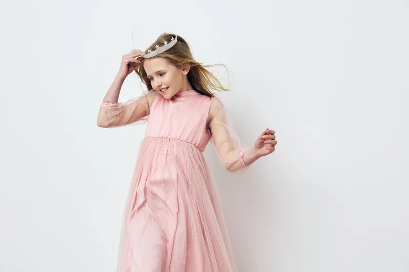 Cheerful young girl in a pink dress with a crown, expressing joy and elegance against a light background, perfect for children s fashion and celebrations