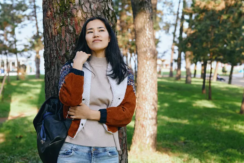 Woman leaning against a tree in a sunny park, showcasing casual outdoor style and relaxed mood among greenery and daylight in a candid lifestyle scene