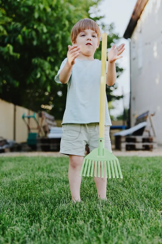Charming boy playing outdoors with a colorful rake, enjoying a sunny day, while expressing curiosity and joy in a vibrant green garden.