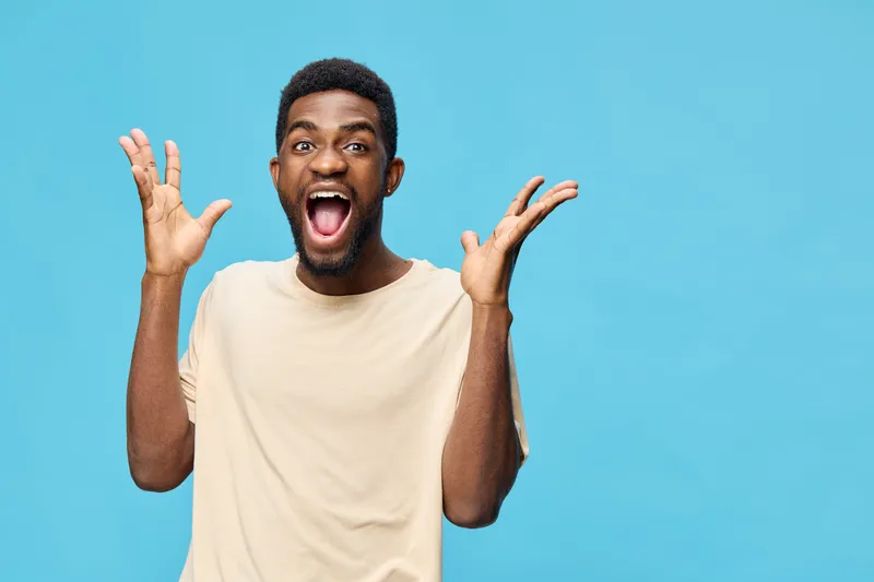 Excited young African American man expressing joy with hands raised against a blue background, showcasing vibrant emotions and a casual outfit