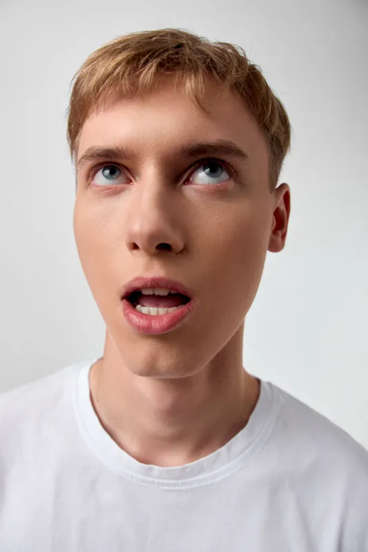 Close up of a surprised young man looking upward, wide eyes and open mouth, wearing a white tee, studio lighting, capturing curiosity and a spontaneous reaction