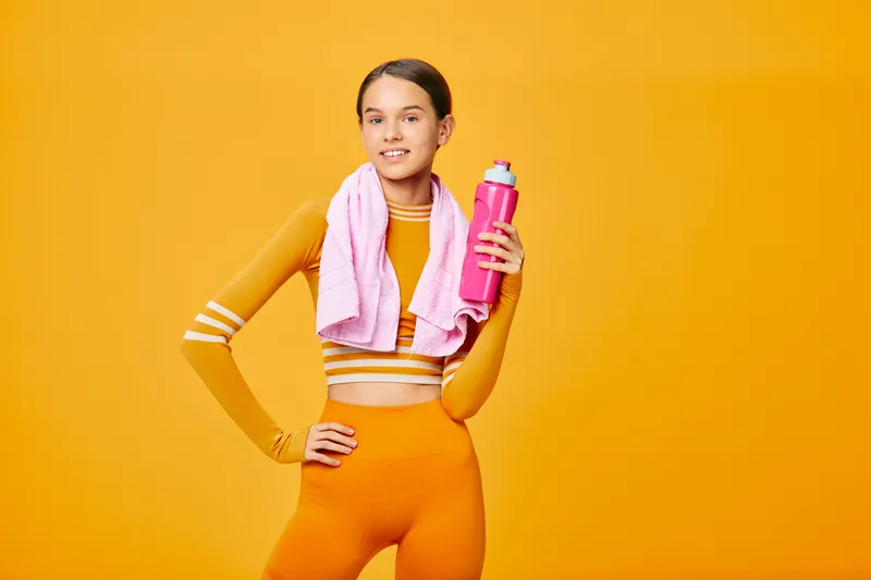 Woman fitness athlete in bright orange athletic wear with a pink water bottle and towel around her neck, posing against a bold orange backdrop for an energetic workout mood.