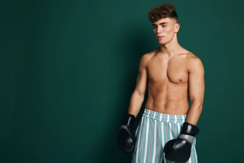 Young man with boxing gloves and striped shorts standing against green background, shirtless athlete preparing for training or workout session in studio environment.