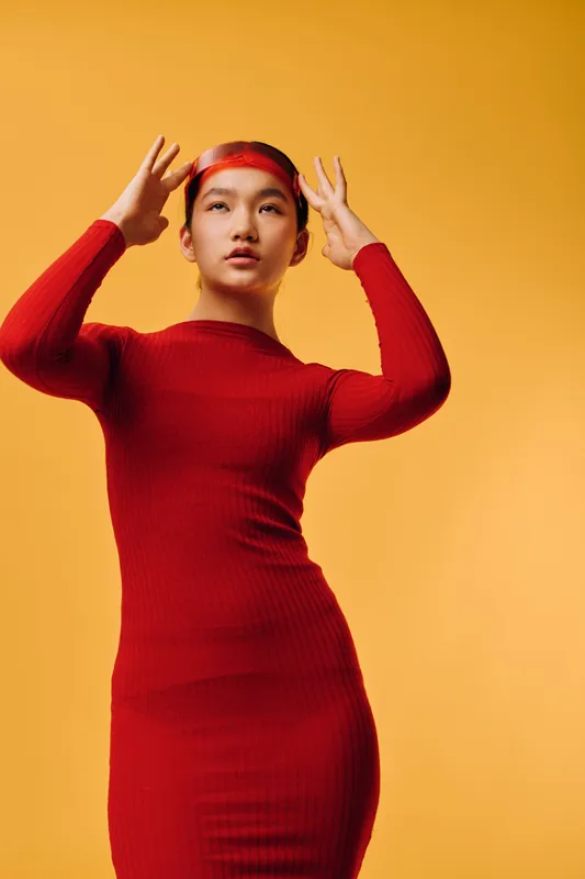 Young woman in red dress and headband posing on yellow background. Fashion model with long hair standing and adjusting accessory for style and beauty portrait.