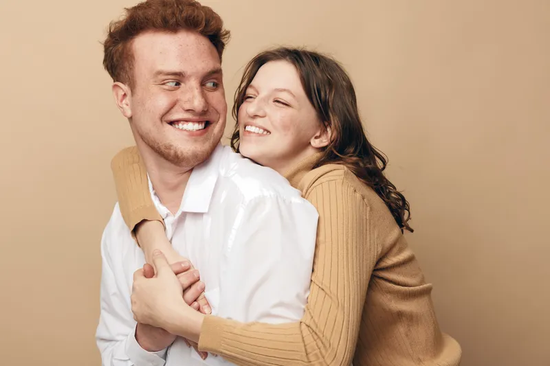 Happy young couple embraces and smiles warmly against beige background. Casual clothing and natural expressions create a cozy, loving atmosphere of friendship and affection.