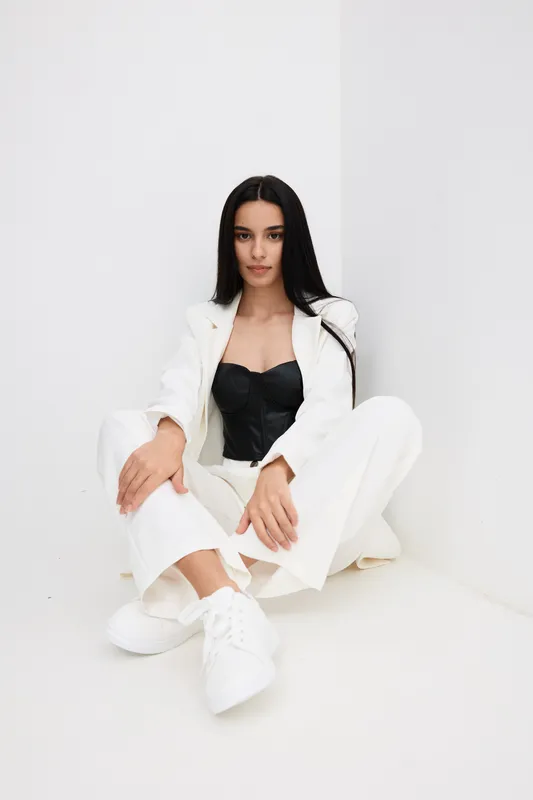Young woman in white suit and black corset sitting on the floor with relaxed posture in a minimalistic white room, stylish modern fashion look.