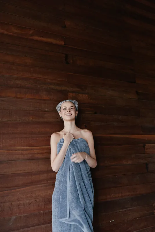 smiling woman wrapped in towel stands against dark wooden wall, hair wrapped with towel, enjoying spa relaxation and calm atmosphere indoors