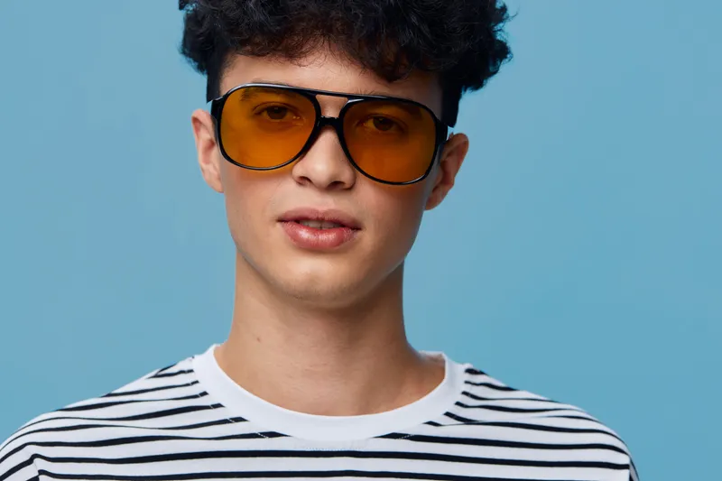 Young man wearing trendy orange sunglasses and striped shirt poses confidently against a blue background, showcasing modern casual style and cool attitude.