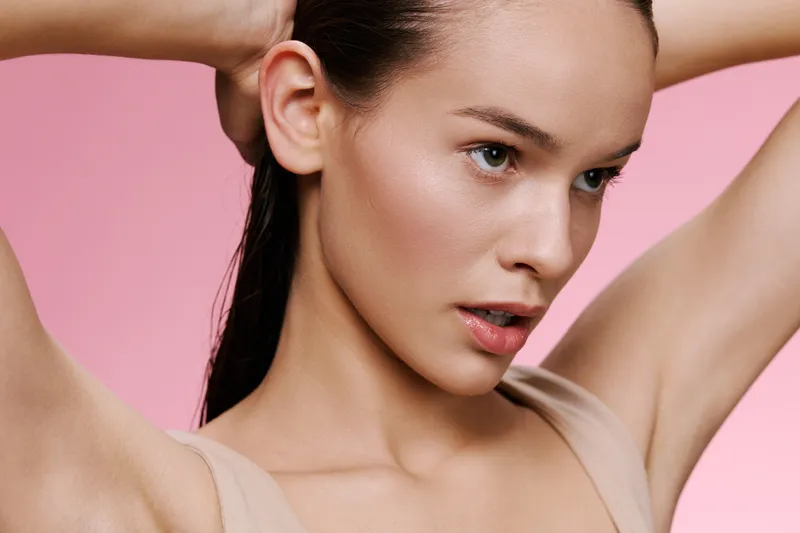 Young woman with long hair pulling it back against a pink background, showcasing a natural beauty and serene expression The model wears minimalistic, neutral toned clothing
