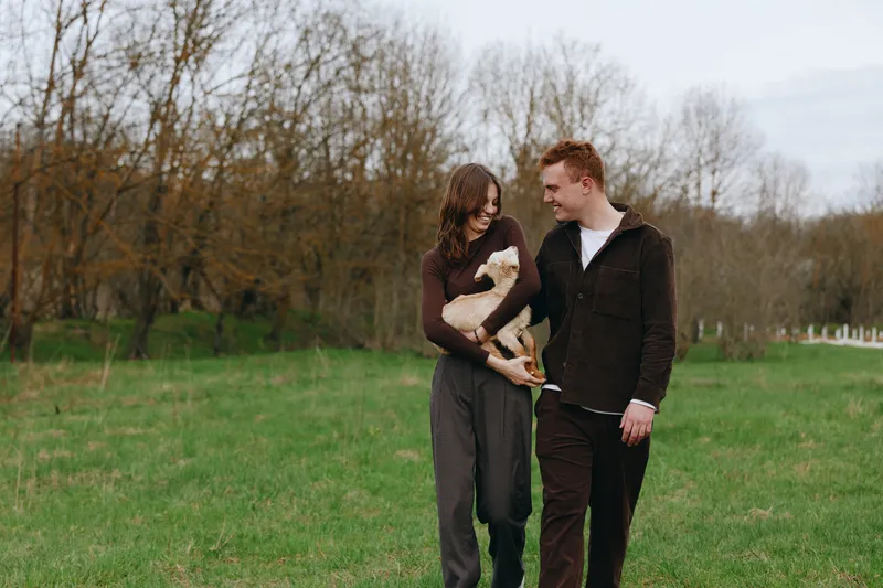 Cheerful couple outdoors with a playful goat kid, strolling on a sunlit field, gentle interaction, casual attire, natural colors, calm mood, soft breeze, rustic backdrop, candid moment of connection