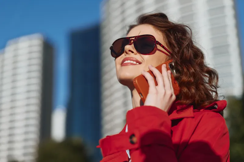 Young woman in a bright red jacket talks on a vivid orange smartphone in a sunny urban setting, wearing oversized sunglasses and a wide smile as tall modern buildings frame the background.