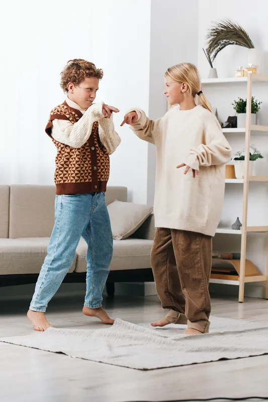 Two children dance indoors, pointing playfully at each other with bright smiles. They stand barefoot on a soft rug in a cozy, sunlit living room, capturing candid, joyful movement.