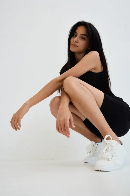 young woman squatting in black dress and white sneakers, long dark hair, minimalistic studio background, casual confident pose, natural lighting, contemporary style