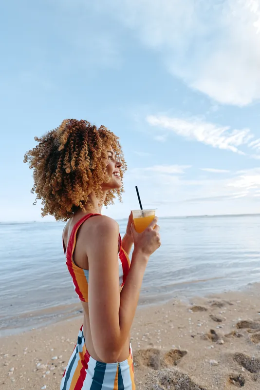 Young woman with curly hair stands on a sandy beach, sipping orange juice from a cup with a straw, enjoying a sunny day by the water.