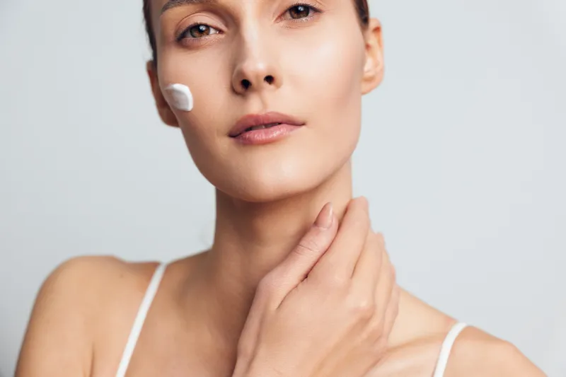 Young woman with clean skin applies moisturizing cream on face touching neck looking at camera with calm expression isolated on light background in skincare beauty concept.