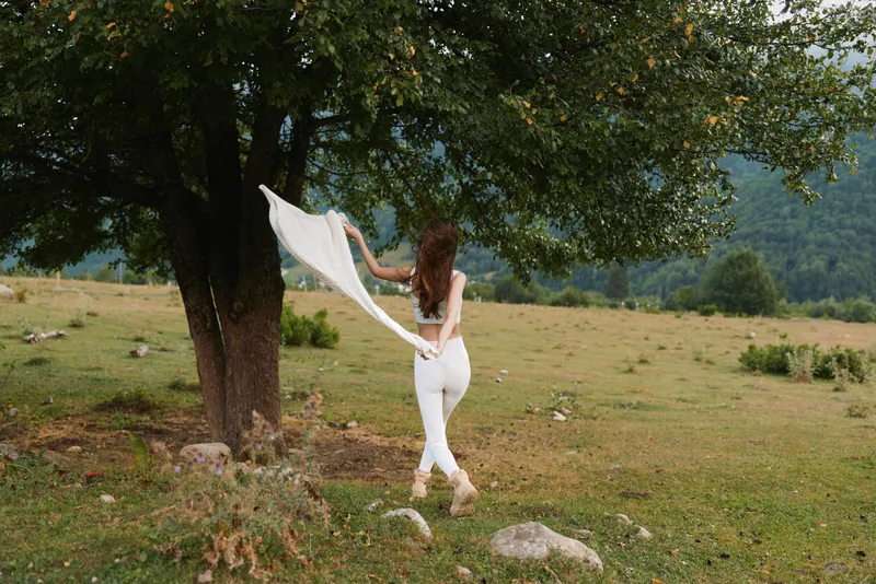 A woman dances outdoors in a sunlit meadow, white scarf fluttering as she spins beside a tree. A moment of carefree movement and natural, everyday joy.