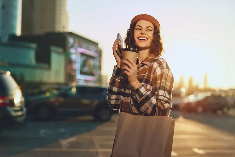Woman holding coffee and shopping bag, smiling in urban street at golden hour, candid moment with sunlight, authenticity, mindful living and emotional storytelling in modern city lifestyle.