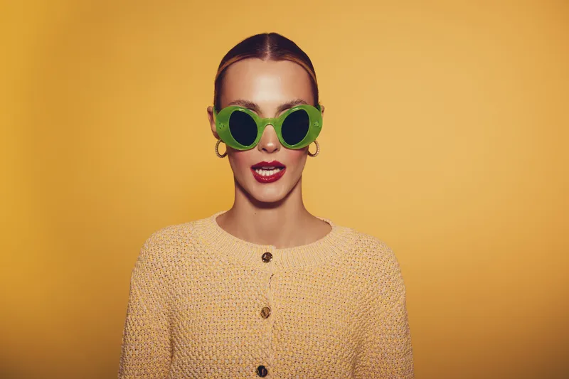 Young woman with slicked hair wearing green round sunglasses and yellow knitted sweater poses against bright yellow background. Fashion concept with bold accessories and confident expression.