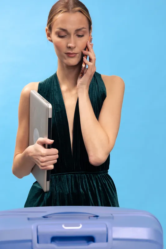 Young woman in elegant dress holds laptop and talks on phone while standing behind suitcase. Concept of business travel, communication, and modern professional lifestyle in studio setting.