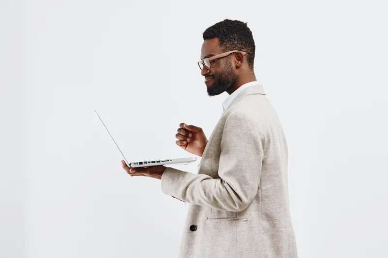 Confident young man with dark skin, wearing beige striped suit, holding and looking at a laptop, smiling, casual posture, isolated on white background. People lifestyle concept, professional or casual