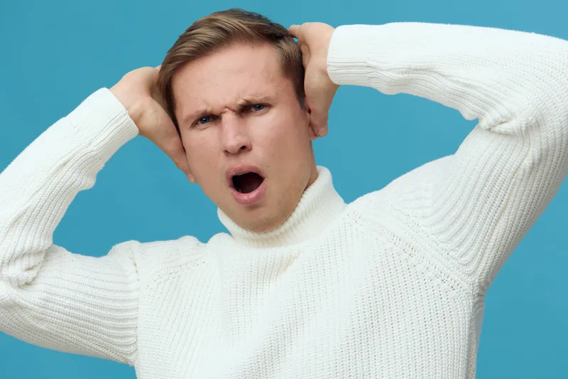 Frustrated young man with brown hair wearing white sweater expressive upset face holding hands on head isolated on blue background studio portrait. People emotion concept