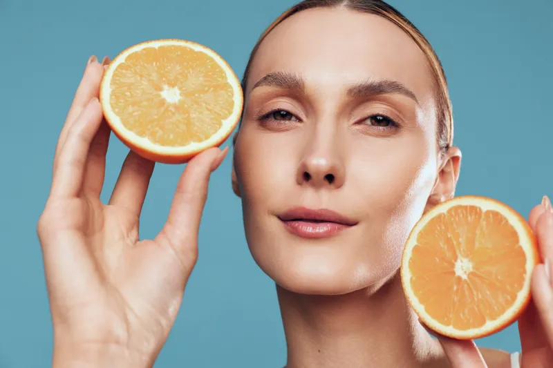 A radiant woman holds orange halves near her face, highlighting fresh citrus and luminous skin against a vibrant blue backdrop, creating a bold health and beauty concept for skincare campaigns.
