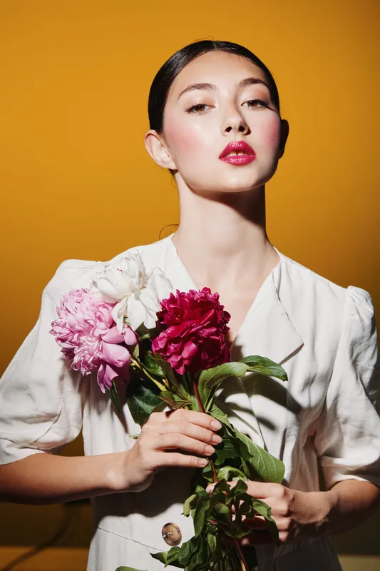 Young woman with glossy makeup and burgundy lips holding a bouquet of peonies in a white top against a bright yellow background, fashion beauty creative concept.