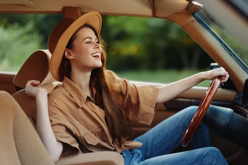 Cheerful young woman driving classic car, wearing stylish hat and casual outfit, showcasing happiness and freedom in a sunny natural setting.
