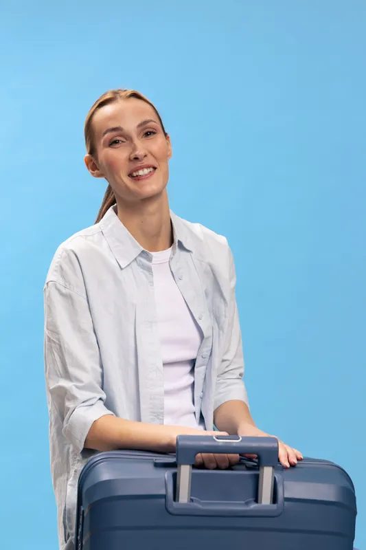 Smiling woman in casual shirt holds blue suitcase against blue background. Concept of travel, vacation, packing, and preparation for trip with positive mood and casual style.