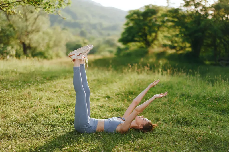 Young woman practicing yoga outdoors in a green field, embodying tranquility and fitness with legs raised and arms extended, dressed in comfortable athletic gear