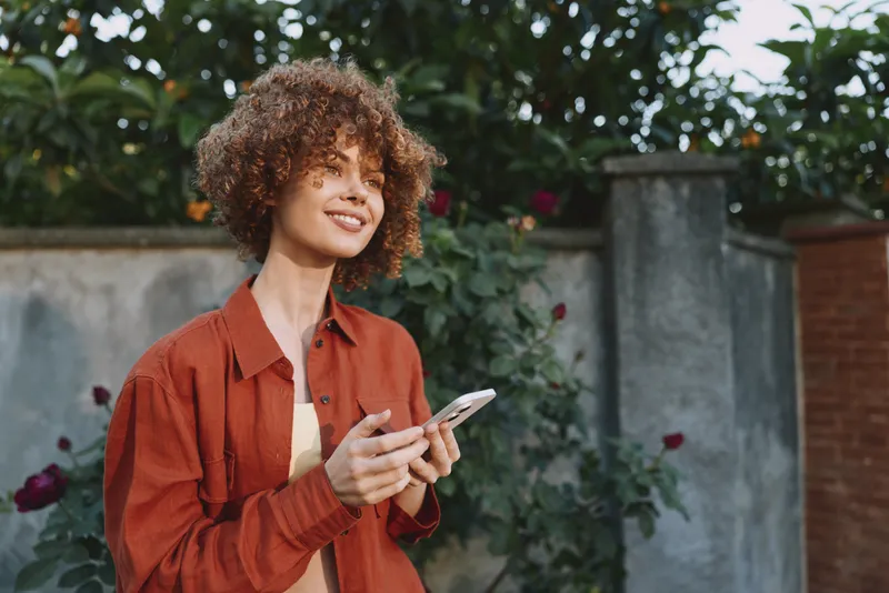 Young woman with curly hair smiles while using a smartphone in a sunny garden, wearing a rust shirt for a casual lifestyle moment