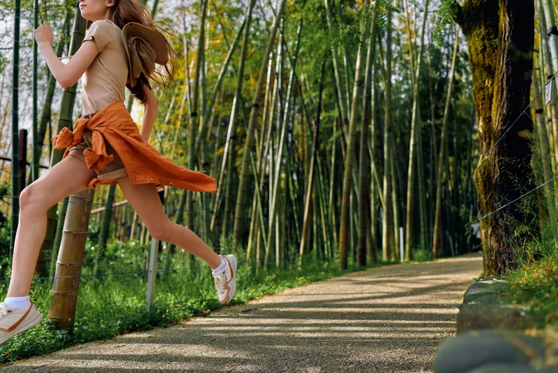 woman jump running bamboo path forest nature sneakers in a lively outdoor scene, young traveler leaping along a sunlit trail with casual skirt, hat and carefree motion through green woods
