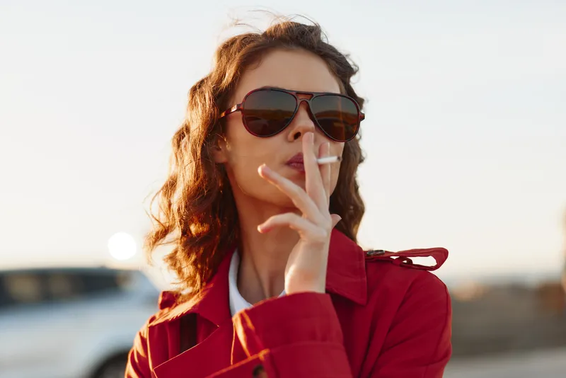 Confident woman in a red jacket wears sunglasses and smokes a cigarette outdoors, capturing a bold street portrait. Theme: smoking, attitude, rebellion.