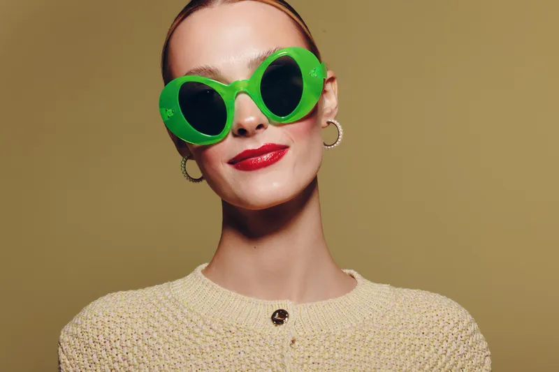 Stylish woman with short hair wearing large green sunglasses and red lipstick smiles confidently against beige background. Fashion concept with bold accessories and modern style.