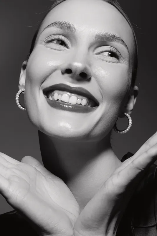 Close-up of a happy caucasian woman with fair skin smiling and posing with hands near face. Studio portrait with earrings, natural makeup, and joyful expression in black and white.