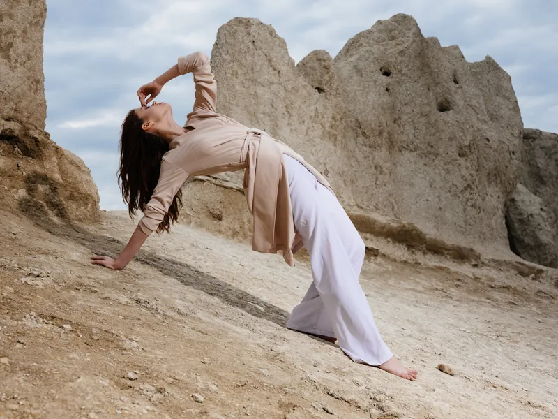 Женщина в светлой одежде стоит на песке рядом со скалами и высокими камнями