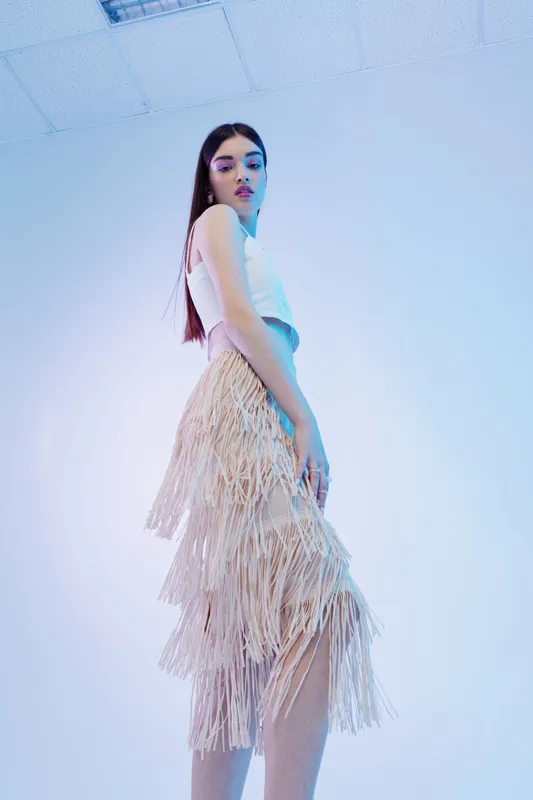 Young woman in a white top and fringed skirt poses in a studio with blue lighting. Concept of beauty, fashion, and modern style for editorial or advertising.