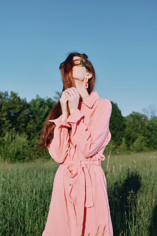 Young woman in summer pink dress standing in a green field, expressing joy and freedom, surrounded by nature under a clear blue sky