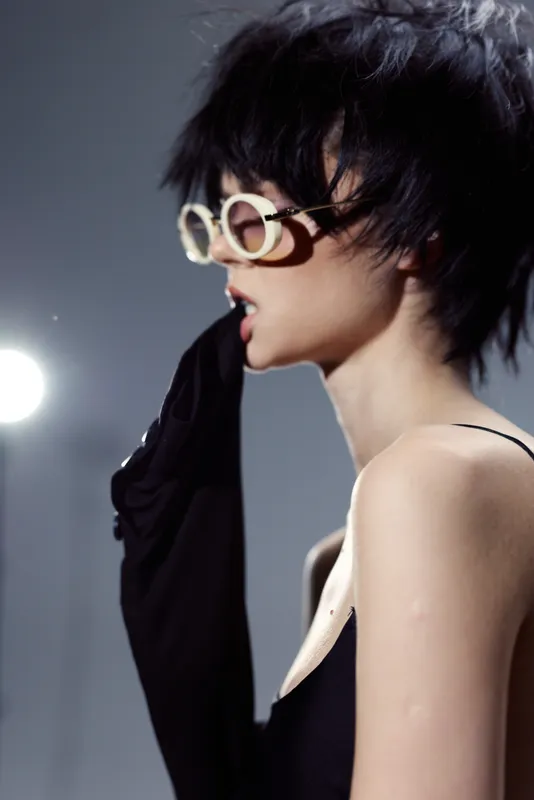 Stylish woman in black outfit biting sleeve wearing round glasses with tousled short hair against gray background.