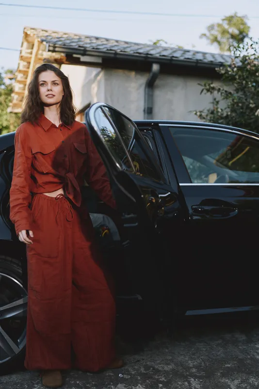 Vibrant woman lifestyle moment with a young woman in a red dress beside a sleek black car, doorway open, relaxed pose, outdoor daytime setting, candid fashion scene