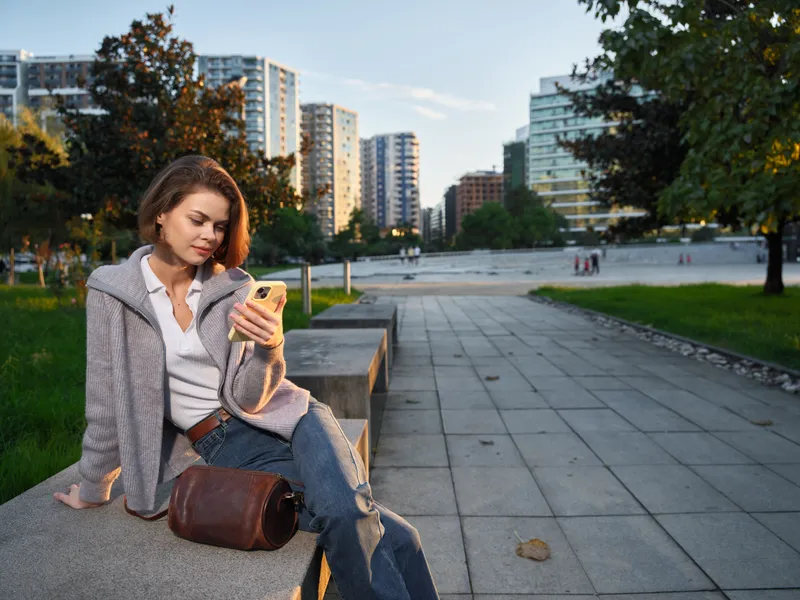 A stylish woman sits on a park bench in a modern city setting, texting on her smartphone. She looks relaxed and focused, enjoying a calm moment amid urban activity.