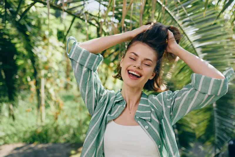 Joyful woman outdoors, laughing with her hands in her hair, wearing a green striped shirt. A bright candid moment captured in natural light.