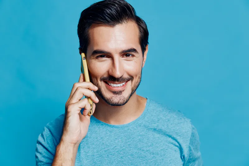 Smiling man in a blue t-shirt holding a smartphone, looking cheerful and engaged while communicating. Isolated on a solid blue background, portrait shot.