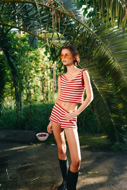 A stylish woman stands in a lush tropical garden wearing a red striped crop top and matching shorts with tall boots, posing confidently under swaying palm fronds and dappled sunlight.