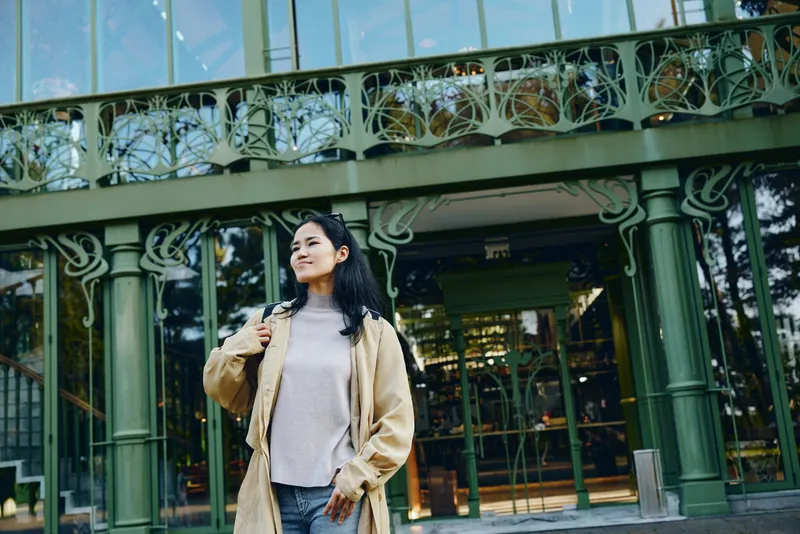 Woman wearing a beige coat on a city street in front of a green storefront illustrates urban fashion, outdoor travel vibe, modern architecture and storefront detail