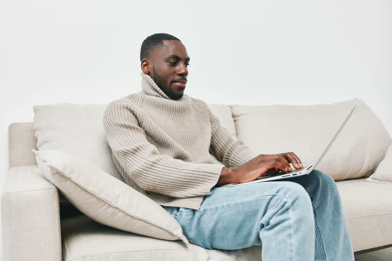 Young Black man working on a laptop at home, wearing a cozy knitted sweater, showcasing focus and comfort in a minimalistic setting