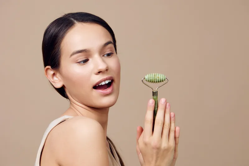 Young woman with fair skin holds a jade roller, expressing joy in a neutral background, embodying beauty and wellness concepts for skincare enthusiasts