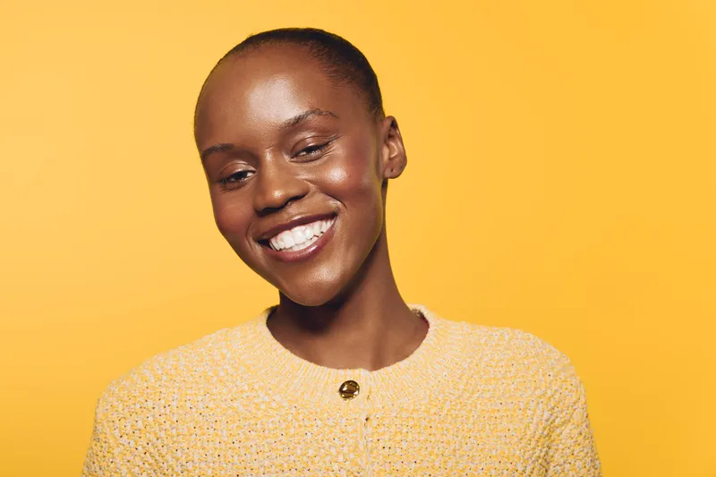 Smiling African woman with dark skin wearing a textured yellow sweater against a yellow background. Concept of happiness, beauty, and confidence in natural portrait photography.