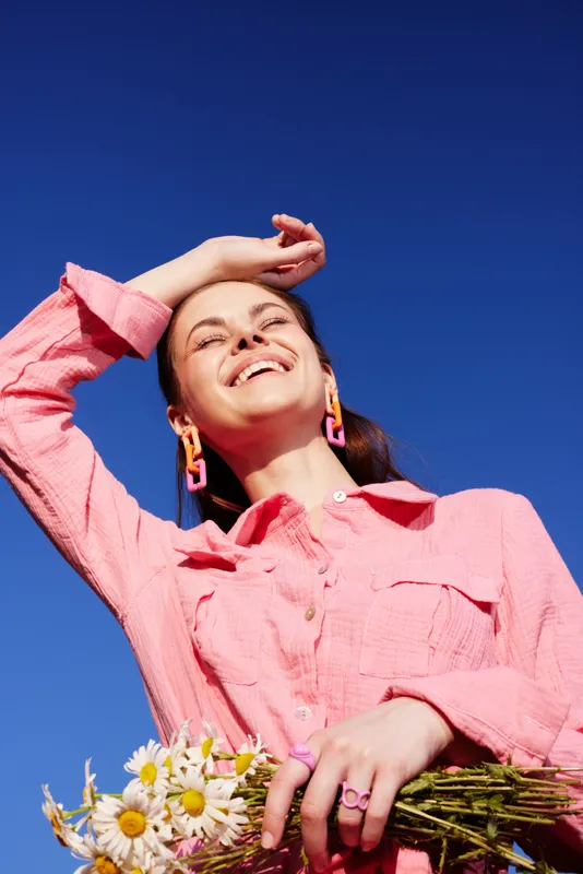 Happy young woman with a floral bouquet, wearing a pink shirt and bold earrings against a vibrant blue sky, conveying joy and positivity in a seasonal spring setting