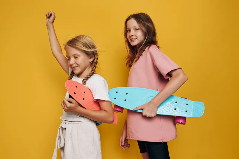 Two happy girls holding colorful skateboards pose against a yellow background. One girl raises her arm in excitement, both smiling. Concept for childhood fun and play.